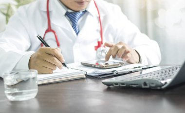 Doktor not defterine bilgi yazıyor ve sağlıklı bir şekilde bakabilmek için telefona bakıyor.