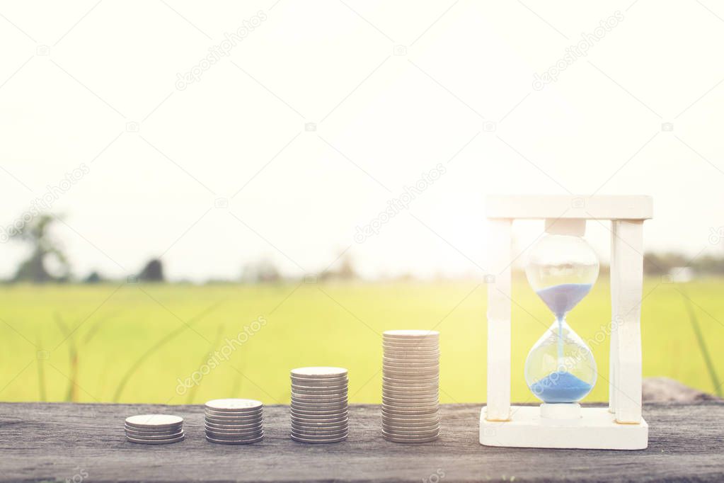 Stacked of money coin and hourglass on wood table, time to save money concept.