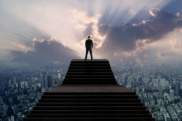 Успех и концепция лидера, бизнесмен в костюме, стоящий на вершине лестницы и смотрящий на город с солнечным светом
 