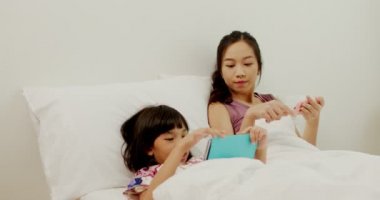 İki Asyalı kız yatakta çocuk telefonu çalar, Taylandlı kız kardeş uyumadan önce akıllı telefonlara bakar ve başkalarına sataşır.