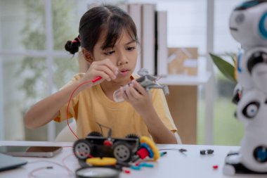 Asian girl focused on connecting robot circuit with wires and tools at home, concept of STEM education, DIY robotics workshop, hands-on engineering, and creative technology learning for children.