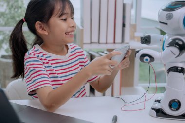 Asian girl smiling while controlling robot with smartphone at home, concept of STEM education, artificial intelligence, coding, and future technology learning for kids.