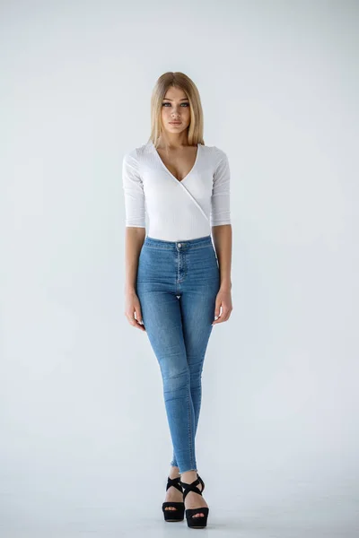 白いシャツと白い背景の上に立ってブルー ジーンズで素敵なブロンドの女の子のスタジオ撮影 — ストック写真