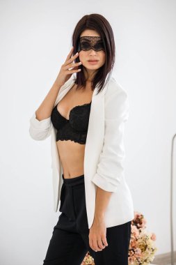 Stüdyo portre siyah pantolon, beyaz ceket ve siyah sütyen evde çiçek kokulu arka plan üzerinde duran güzel esmer kadının yüzünde siyah maske ile 