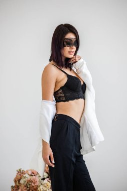 Stüdyo portre siyah pantolon, beyaz ceket ve siyah sütyen evde çiçek kokulu arka plan üzerinde duran güzel esmer kadının yüzünde siyah maske ile 