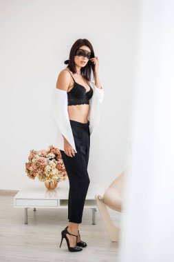 Stüdyo portre siyah pantolon, beyaz ceket ve siyah sütyen evde çiçek kokulu arka plan üzerinde duran güzel esmer kadının yüzünde siyah maske ile 