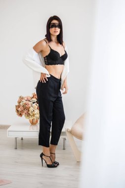 Stüdyo portre siyah pantolon, beyaz ceket ve siyah sütyen evde çiçek kokulu arka plan üzerinde duran güzel esmer kadının yüzünde siyah maske ile 