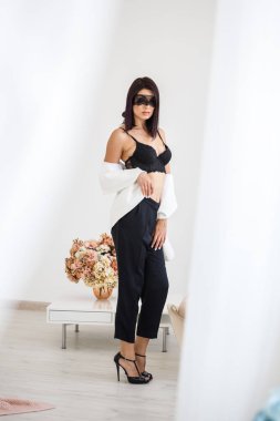 Stüdyo portre siyah pantolon, beyaz ceket ve siyah sütyen evde çiçek kokulu arka plan üzerinde duran güzel esmer kadının yüzünde siyah maske ile 