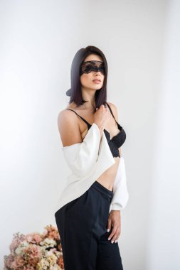 Stüdyo portre siyah pantolon, beyaz ceket ve siyah sütyen evde çiçek kokulu arka plan üzerinde duran güzel esmer kadının yüzünde siyah maske ile 