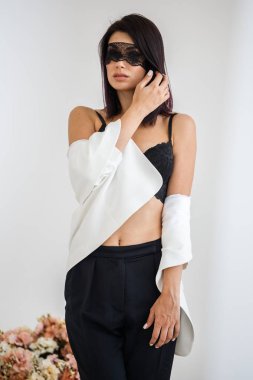 Stüdyo portre siyah pantolon, beyaz ceket ve siyah sütyen evde çiçek kokulu arka plan üzerinde duran güzel esmer kadının yüzünde siyah maske ile 