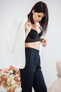 Güzel esmer kadın giyen siyah pantolon, beyaz ceket ve siyah sütyen ayakta, çiçek kokulu arka plan üzerinde ev iç stüdyo portresi 
