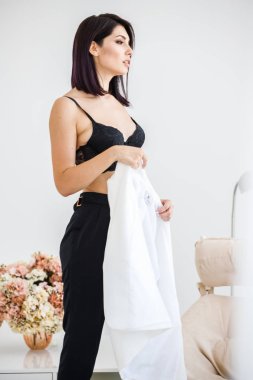 Güzel esmer kadın giyen siyah pantolon, beyaz ceket ve siyah sütyen ayakta, çiçek kokulu arka plan üzerinde ev iç stüdyo portresi 