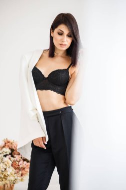 Güzel esmer kadın giyen siyah pantolon, beyaz ceket ve siyah sütyen ayakta, çiçek kokulu arka plan üzerinde ev iç stüdyo portresi 