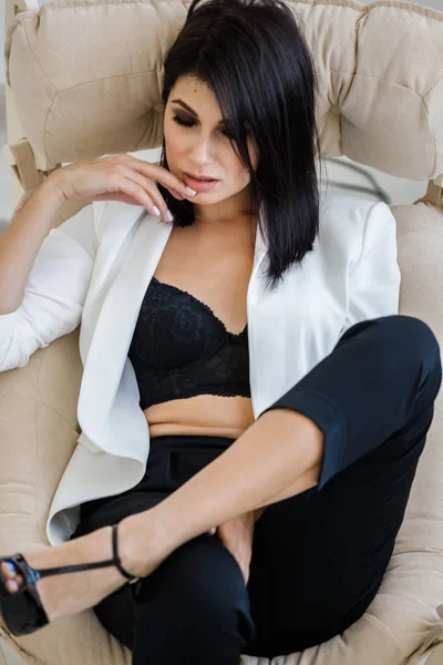 在家里的扶手椅上穿着黑色裤子 白色夹克和黑色胸罩的漂亮黑发女人的工作室肖像 — 图库照片