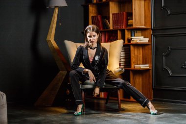 Pembe tişört ve koltuk ile kitaplık arka plan üzerine oturmuş siyah takım elbise giyen güzel esmer kadın portresi 