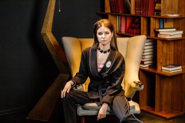 Pembe tişört ve koltuk ile kitaplık arka plan üzerine oturmuş siyah takım elbise giyen güzel esmer kadın portresi 