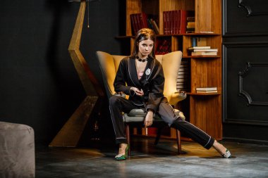 Pembe tişört ve koltuk ile kitaplık arka plan üzerine oturmuş siyah takım elbise giyen güzel esmer kadın portresi 