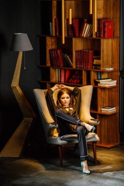 Pembe tişört ve koltuk ile kitaplık arka plan üzerine oturmuş siyah takım elbise giyen güzel esmer kadın portresi 