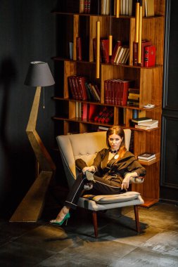 Pembe tişört ve koltuk ile kitaplık arka plan üzerine oturmuş siyah takım elbise giyen güzel esmer kadın portresi 