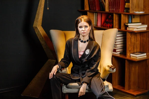 Pembe tişört ve koltuk ile kitaplık arka plan üzerine oturmuş siyah takım elbise giyen güzel esmer kadın portresi 