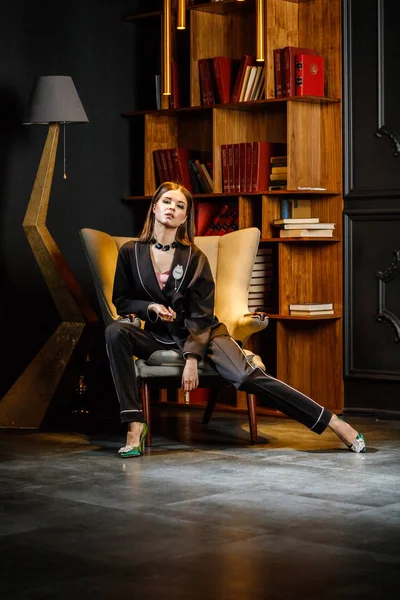 Pembe tişört ve koltuk ile kitaplık arka plan üzerine oturmuş siyah takım elbise giyen güzel esmer kadın portresi 