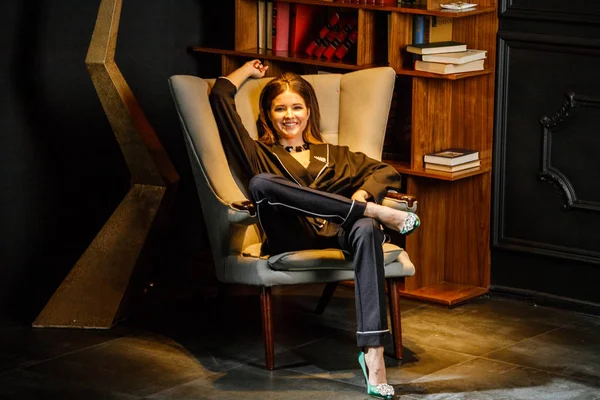 Pembe tişört ve koltuk ile kitaplık arka plan üzerine oturmuş siyah takım elbise giyen güzel esmer kadın portresi 