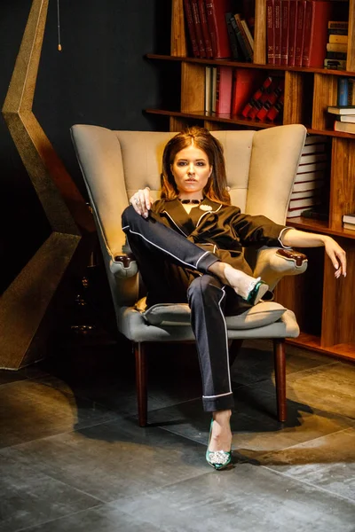 Pembe tişört ve koltuk ile kitaplık arka plan üzerine oturmuş siyah takım elbise giyen güzel esmer kadın portresi 