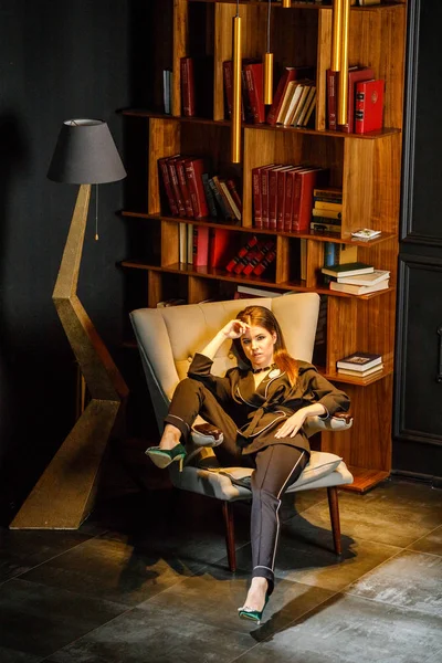 Pembe tişört ve koltuk ile kitaplık arka plan üzerine oturmuş siyah takım elbise giyen güzel esmer kadın portresi 