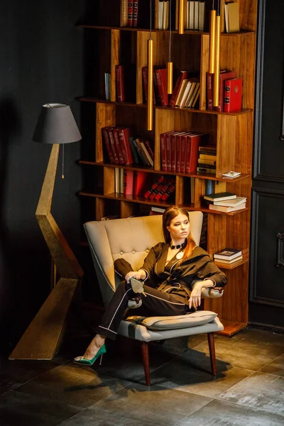 Pembe tişört ve koltuk ile kitaplık arka plan üzerine oturmuş siyah takım elbise giyen güzel esmer kadın portresi 