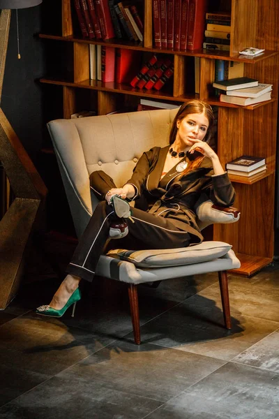 Pembe tişört ve koltuk ile kitaplık arka plan üzerine oturmuş siyah takım elbise giyen güzel esmer kadın portresi 