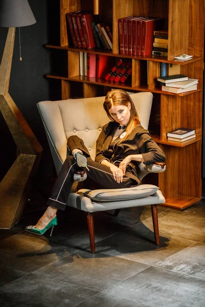 Pembe tişört ve koltuk ile kitaplık arka plan üzerine oturmuş siyah takım elbise giyen güzel esmer kadın portresi 