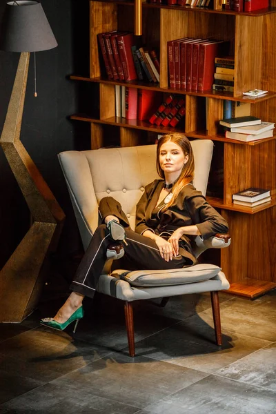 Pembe tişört ve koltuk ile kitaplık arka plan üzerine oturmuş siyah takım elbise giyen güzel esmer kadın portresi 