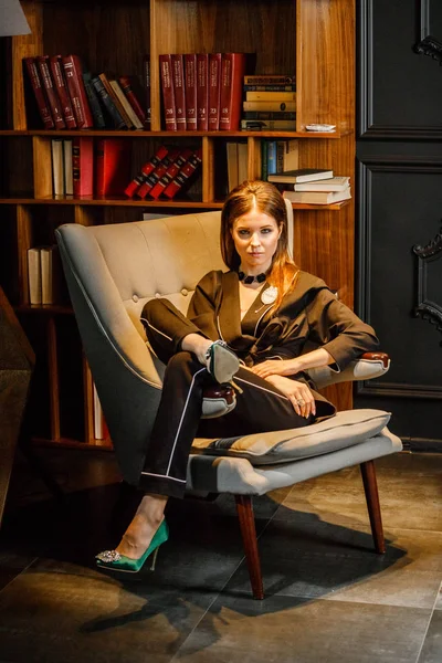 Pembe tişört ve koltuk ile kitaplık arka plan üzerine oturmuş siyah takım elbise giyen güzel esmer kadın portresi 