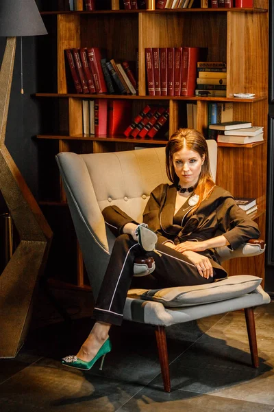 Pembe tişört ve koltuk ile kitaplık arka plan üzerine oturmuş siyah takım elbise giyen güzel esmer kadın portresi 