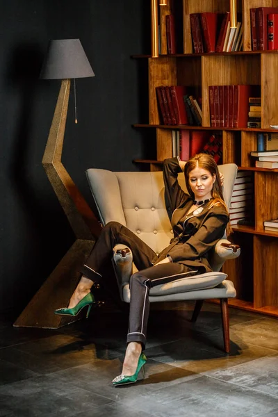 Pembe tişört ve koltuk ile kitaplık arka plan üzerine oturmuş siyah takım elbise giyen güzel esmer kadın portresi 