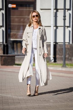 Açık bej ceket, beyaz gömlek ve beyaz pantolon giyiyor, bulanık arka planda bina ile sokakta duran güzellik kadın portresi