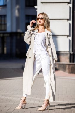 Açık bej ceket, beyaz gömlek ve beyaz pantolon giyiyor, bulanık arka planda bina ile sokakta duran güzellik kadın portresi