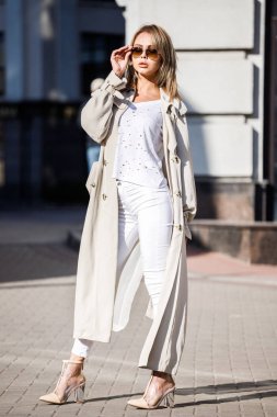 Açık bej ceket, beyaz gömlek ve beyaz pantolon giyiyor, bulanık arka planda bina ile sokakta duran güzellik kadın portresi