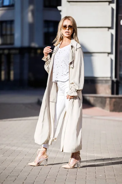 Açık bej ceket, beyaz gömlek ve beyaz pantolon giyiyor, bulanık arka planda bina ile sokakta duran güzellik kadın portresi