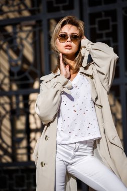 Açık bej ceket, beyaz gömlek ve beyaz pantolon giyiyor, bulanık arka planda bina ile sokakta duran güzellik kadın portresi