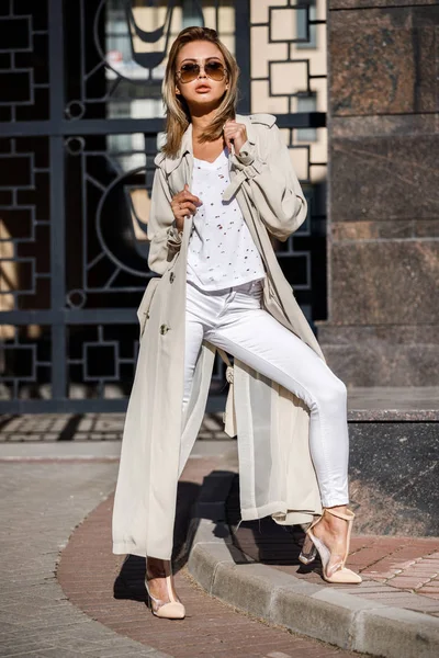 Açık bej ceket, beyaz gömlek ve beyaz pantolon giyiyor, bulanık arka planda bina ile sokakta duran güzellik kadın portresi