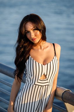 Çizgili siyah beyaz elbiseli güzel genç esmer kadın açık bir kadeh. Günbatımı sırasında iskelede duran kıvırcık kadın deniz arka plan bulanık 