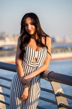 Çizgili siyah beyaz elbiseli güzel genç esmer kadın açık bir kadeh. Güneş ışığı, şehir dolgu üzerinde duran kıvırcık kadın şehir görünümü arka plan bulanık 