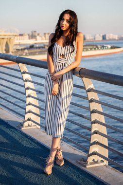 Çizgili siyah beyaz elbiseli güzel genç esmer kadın açık bir kadeh. Güneş ışığı, şehir dolgu üzerinde duran kıvırcık kadın şehir görünümü arka plan bulanık 