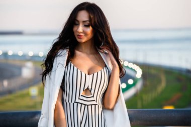 Çizgili siyah beyaz elbiseli güzel genç esmer kadın açık bir kadeh. Güneş ışığı, şehir dolgu üzerinde duran kıvırcık kadın şehir görünümü arka plan bulanık 