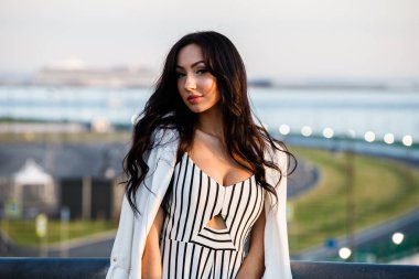 Çizgili siyah beyaz elbiseli güzel genç esmer kadın açık bir kadeh. Güneş ışığı, şehir dolgu üzerinde duran kıvırcık kadın şehir görünümü arka plan bulanık 