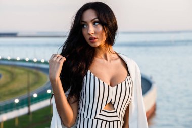 Çizgili siyah beyaz elbiseli güzel genç esmer kadın açık bir kadeh. Güneş ışığı, şehir dolgu üzerinde duran kıvırcık kadın şehir görünümü arka plan bulanık 