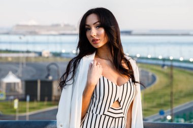 Çizgili siyah beyaz elbiseli güzel genç esmer kadın açık bir kadeh. Güneş ışığı, şehir dolgu üzerinde duran kıvırcık kadın şehir görünümü arka plan bulanık 