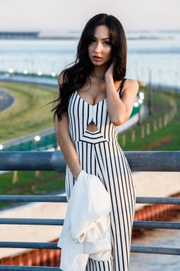 Çizgili siyah beyaz elbiseli güzel genç esmer kadın açık bir kadeh. Güneş ışığı, şehir dolgu üzerinde duran kıvırcık kadın şehir görünümü arka plan bulanık 