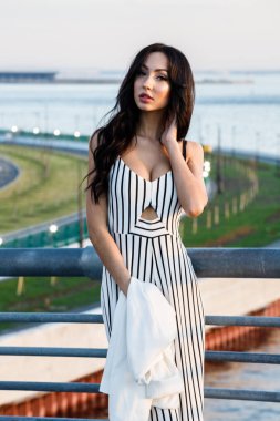 Çizgili siyah beyaz elbiseli güzel genç esmer kadın açık bir kadeh. Güneş ışığı, şehir dolgu üzerinde duran kıvırcık kadın şehir görünümü arka plan bulanık 